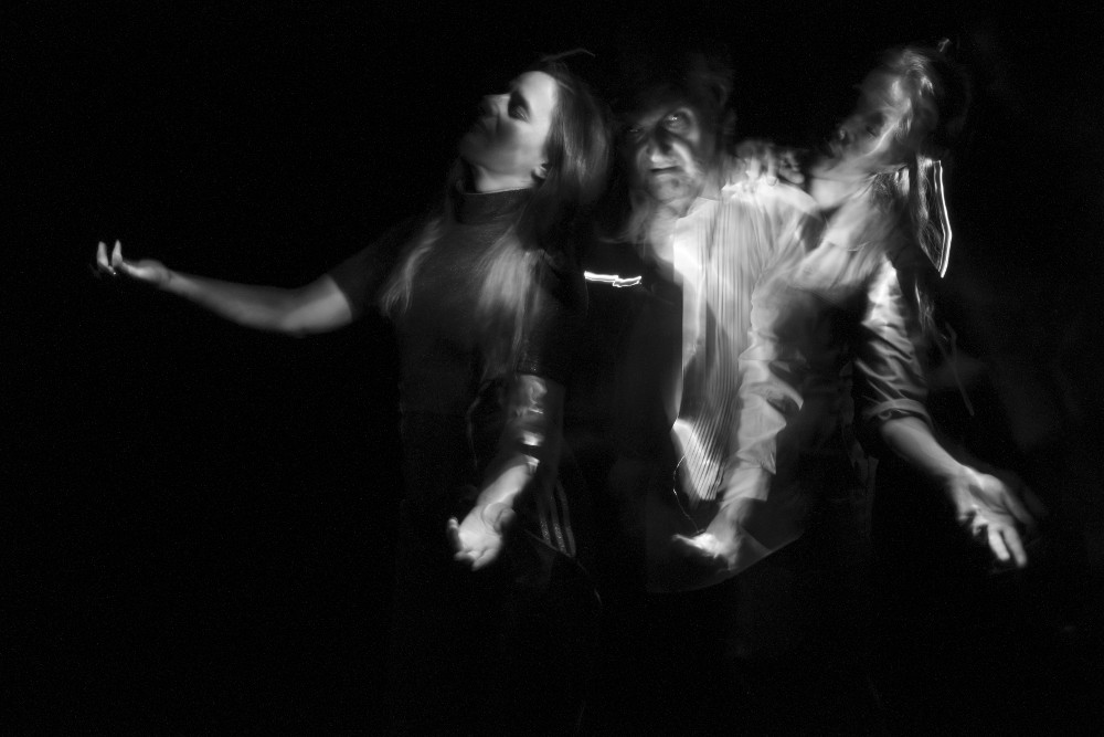 Vom Dunkeln VIII (Selbstportrait mit Rachael Dichter und Sherwood Chen) (2022) von Gerald Pirner ist eine Schwarz-Weiß-Fotografie im Querformat, die mithilfe der Lightpainting-Technik erstellt wurde.Zu sehen sind drei Personen vom Kopf bis zur Hüfte vor einem schwarzen Hintergrund. Sie stehen dicht an dicht und leicht hintereinander. Alle haben den linken Arm mit offenen Handflächen nach vorne gerichtet. Die erste Person (Rachael Dichter) links im Bild hat zusätzlich den rechten Arm nach oben gerichtet. Ihre Augen sind geschlossen und ihr Gesicht ist nach links gerichtet. Die Person in der Mitte (Gerald Pirner) hat die Augen geöffnet. Die Person rechts (Sherwood Chen) hat ihre Hand auf der Schulter der mittleren Person. Sie hat die Augen geschlossen und ihr Gesicht ist halb im Dunkeln. Arianna Waldner Bingemer Vom Dunkeln VIII (Selbstportrait mit Rachael Dichter und Sherwood Chen) (2022) von Gerald Pirner ist eine Schwarz-Weiß-Fotografie im Querformat, die mithilfe der Lightpainting-Technik erstellt wurde.Zu sehen sind drei Personen vom Kopf bis zur Hüfte vor einem schwarzen Hintergrund. Sie stehen dicht an dicht und leicht hintereinander. Alle haben den linken Arm mit offenen Handflächen nach vorne gerichtet. Die erste Person (Rachael Dichter) links im Bild hat zusätzlich den rechten Arm nach oben gerichtet. Ihre Augen sind geschlossen und ihr Gesicht ist nach links gerichtet. Die Person in der Mitte (Gerald Pirner) hat die Augen geöffnet. Die Person rechts (Sherwood Chen) hat ihre Hand auf der Schulter der mittleren Person. Sie hat die Augen geschlossen und ihr Gesicht ist halb im Dunkeln. Arianna Waldner Bingemer