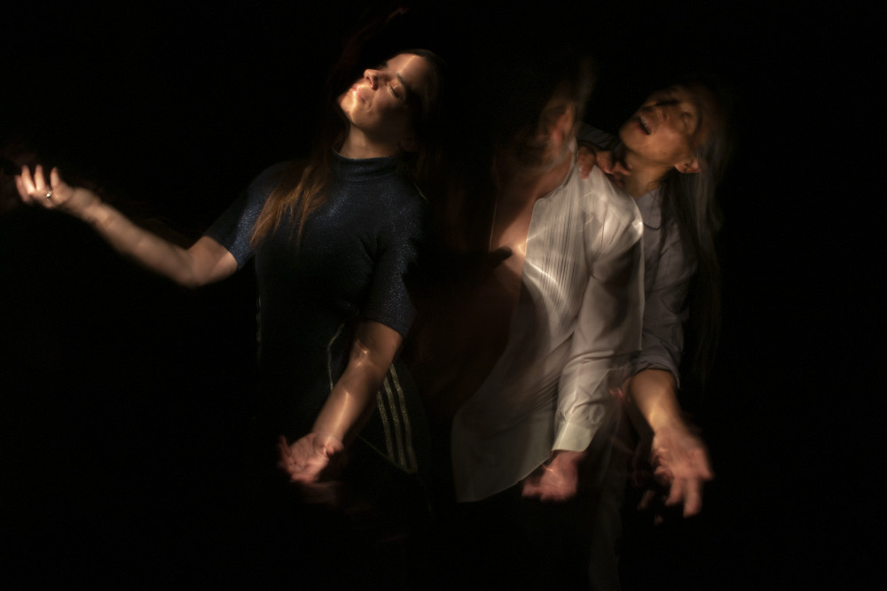 Vom Dunkeln IX (Selbstportrait mit Rachael Dichter und Sherwood Chen), 2022 von Gerald Pirner ist eine Farbfotografie im Querformat, die mithilfe der Lightpainting-Technik erstellt wurde. Zu sehen sind drei Personen vom Kopf bis zum Oberschenkel vor einem schwarzen Hintergrund. Sie stehen dicht an dicht und leicht hintereinander. Alle haben den linken Arm mit offenen Handflächen nach vorne gerichtet. Die erste Person (Rachael Dichter) links im Bild hat zusätzlich den rechten Arm nach oben gerichtet. Ihre Augen sind geschlossen und ihr Gesicht ist hell erleuchtet. Das Gesicht der Person in der Mitte (Gerald Pirner) liegt halb im Dunkeln, der Brustbereich ist durch eine Taschenlampe erleuchtet. Die Person rechts (Sherwood Chen) hat ihre Hand auf der Schulter der mittleren Person. Auch ihre Augen sind geschlossen und er Mund ist leicht geöffnet. Arianna Waldner Bingemer Vom Dunkeln IX (Selbstportrait mit Rachael Dichter und Sherwood Chen), 2022 von Gerald Pirner ist eine Farbfotografie im Querformat, die mithilfe der Lightpainting-Technik erstellt wurde. Zu sehen sind drei Personen vom Kopf bis zum Oberschenkel vor einem schwarzen Hintergrund. Sie stehen dicht an dicht und leicht hintereinander. Alle haben den linken Arm mit offenen Handflächen nach vorne gerichtet. Die erste Person (Rachael Dichter) links im Bild hat zusätzlich den rechten Arm nach oben gerichtet. Ihre Augen sind geschlossen und ihr Gesicht ist hell erleuchtet. Das Gesicht der Person in der Mitte (Gerald Pirner) liegt halb im Dunkeln, der Brustbereich ist durch eine Taschenlampe erleuchtet. Die Person rechts (Sherwood Chen) hat ihre Hand auf der Schulter der mittleren Person. Auch ihre Augen sind geschlossen und er Mund ist leicht geöffnet. Arianna Waldner Bingemer
