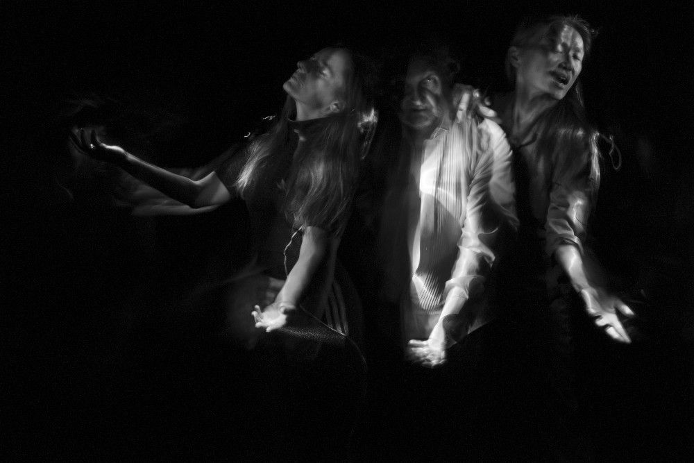 Vom Dunkeln VI (Selbstportrait mit Rachael Dichter und Sherwood Chen), 2022 von Gerald Pirner ist eine Schwarz-Weiß-Fotografie im Querformat, die mithilfe der Lightpainting-Technik erstellt wurde. Zu sehen sind drei Personen vom Kopf bis zur Hüfte vor einem schwarzen Hintergrund. Sie stehen dicht an dicht und leicht hintereinander. Alle haben den linken Arm mit offenen Handflächen nach vorne gerichtet. Die erste Person (Rachael Dichter) links im Bild hat zusätzlich den rechten Arm nach oben gerichtet. Ihre Augen sind geschlossen und ihr Gesicht nach links gerichtet. Das Gesicht der Person in der Mitte (Gerald Pirner) liegt teilweise im Dunkeln, ein Auge ist geöffnet. Die Person rechts (Sherwood Chen) hat ihre Hand auf der Schulter der mittleren Person. Sie ist etwa einen Kopf größer als die anderen, hat die Augen geschlossen und den Kopf nach rechts gerichtet. Arianna Waldner Bingemer Vom Dunkeln VI (Selbstportrait mit Rachael Dichter und Sherwood Chen), 2022 von Gerald Pirner ist eine Schwarz-Weiß-Fotografie im Querformat, die mithilfe der Lightpainting-Technik erstellt wurde. Zu sehen sind drei Personen vom Kopf bis zur Hüfte vor einem schwarzen Hintergrund. Sie stehen dicht an dicht und leicht hintereinander. Alle haben den linken Arm mit offenen Handflächen nach vorne gerichtet. Die erste Person (Rachael Dichter) links im Bild hat zusätzlich den rechten Arm nach oben gerichtet. Ihre Augen sind geschlossen und ihr Gesicht nach links gerichtet. Das Gesicht der Person in der Mitte (Gerald Pirner) liegt teilweise im Dunkeln, ein Auge ist geöffnet. Die Person rechts (Sherwood Chen) hat ihre Hand auf der Schulter der mittleren Person. Sie ist etwa einen Kopf größer als die anderen, hat die Augen geschlossen und den Kopf nach rechts gerichtet. Arianna Waldner Bingemer